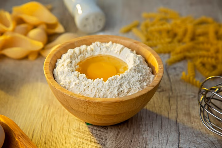 Un bol en bois contenant de la farine et un jaune d'œuf, préparé pour confectionner des pâtes fraîches maison.